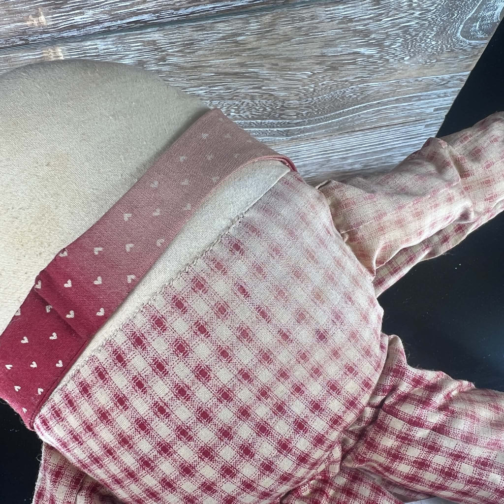 Close-up of the rear clothing on a Humpty Dumpty cloth doll, showing uneven fading across red-and-cream checked fabric and visible stitching along the upper body seam. A cream fabric band separates the checked outfit from the faded heart-patterned collar.