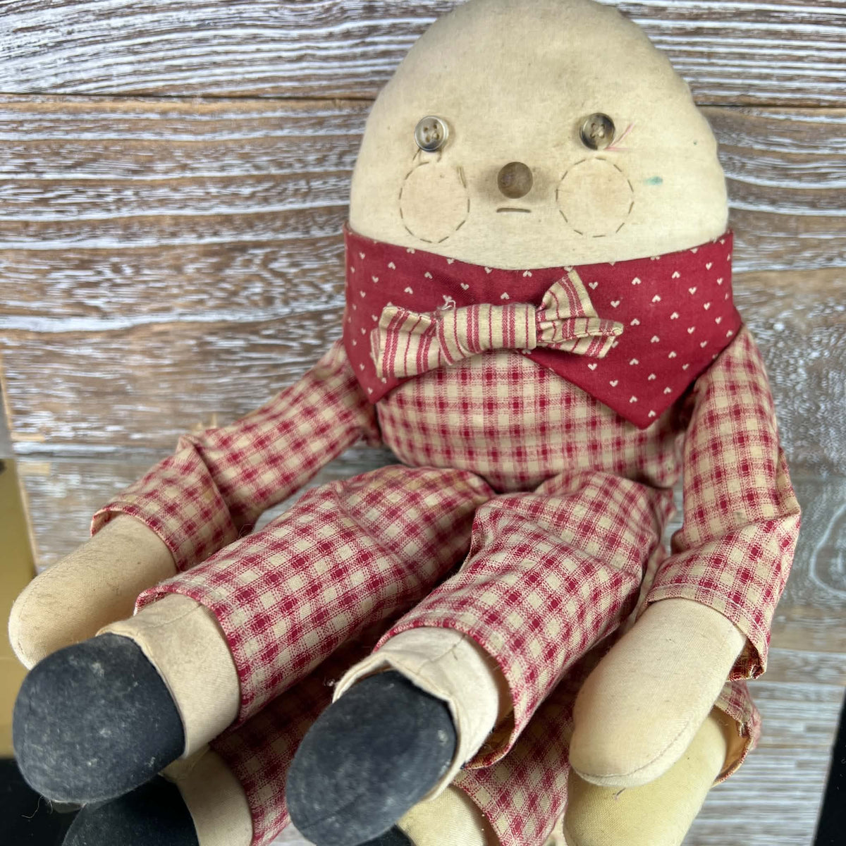 Humpty Dumpty cloth shelf sitter seated against weathered wood, with a cream face, button eyes, stitched circular cheeks and a brown nose. Red checked clothing, a heart-patterned collar and striped bow tie coordinate with cream hands and dark fabric shoes.