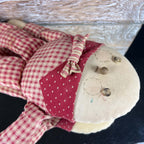 Angled view of a Humpty Dumpty cloth doll resting on a dark surface beside weathered wood. The cream face has button eyes and stitched cheeks, while a red striped bow tie overlaps a heart-print collar above the red-and-cream checked fabric body and limbs.