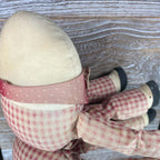 Back view of a Humpty Dumpty cloth shelf sitter showing the rounded cream head and curved seam above a faded red collar patterned with tiny white hearts. Checked fabric covers the body and extended limbs, with dark shoes visible against weathered wood.