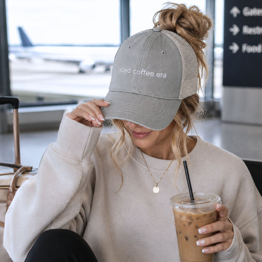 Woman wearing gray and tan mesh back trucker hat with iced coffee era embroidered saying while holding iced coffee at airport terminal