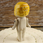 Three-quarter rear view of the vintage Japanese penny doll standing on an ivory crocheted surface. The impressed JAPAN mark, molded curls, age-related paint wear, separate bisque arms, and exposed shoulder fasteners can be seen against a rustic wood background.