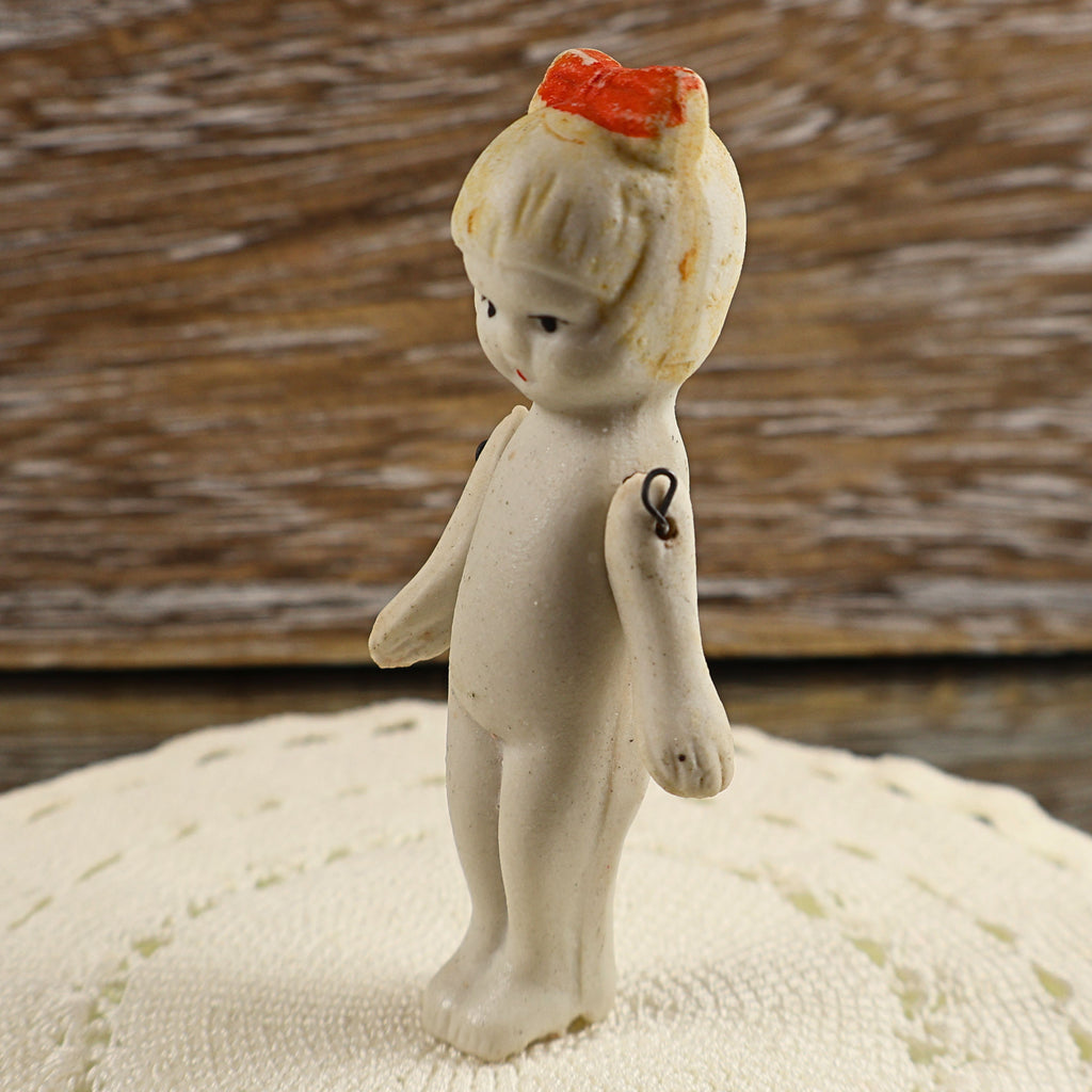 Three-quarter profile of a miniature Japanese bisque girl doll posed on a cream textile. Her painted facial features, small red hair bow, molded wavy hair, slender body, and separate movable arms can be seen, along with darkened metal wire at the shoulder joints.