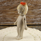 Three-quarter profile of a miniature Japanese bisque girl doll posed on a cream textile. Her painted facial features, small red hair bow, molded wavy hair, slender body, and separate movable arms can be seen, along with darkened metal wire at the shoulder joints.