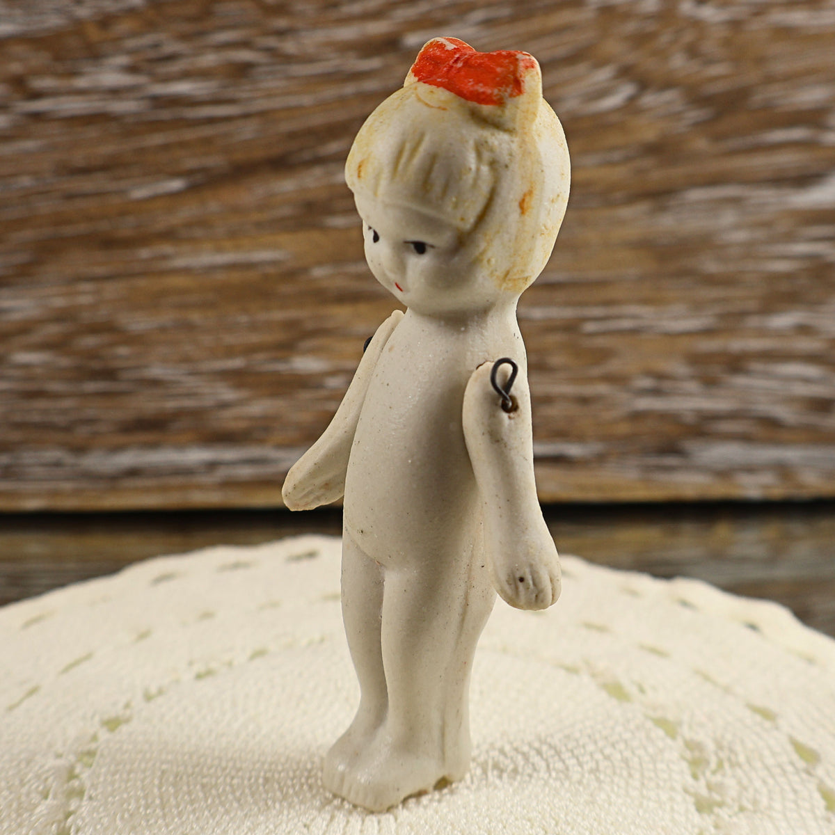 Three-quarter profile of a miniature Japanese bisque girl doll posed on a cream textile. Her painted facial features, small red hair bow, molded wavy hair, slender body, and separate movable arms can be seen, along with darkened metal wire at the shoulder joints.