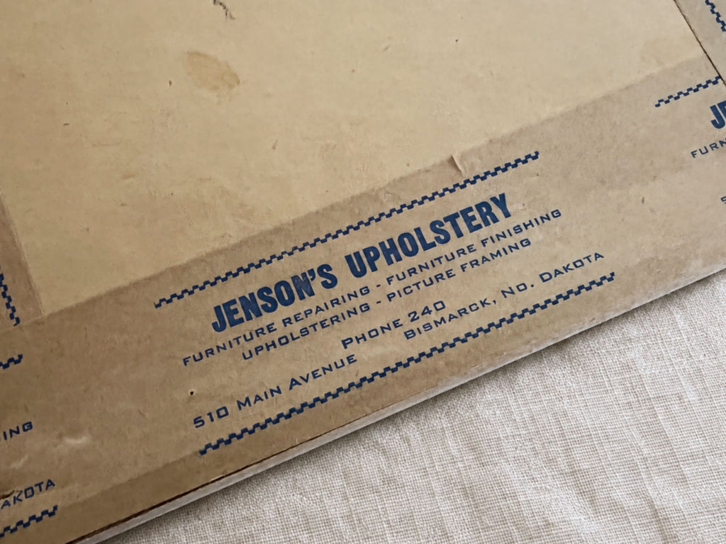 Close-up of blue Jenson’s Upholstery printing on brown framing tape. The label lists furniture repairing, furniture finishing, upholstering, picture framing, Phone 240, and 510 Main Avenue in Bismarck, North Dakota, with backing discoloration visible above.