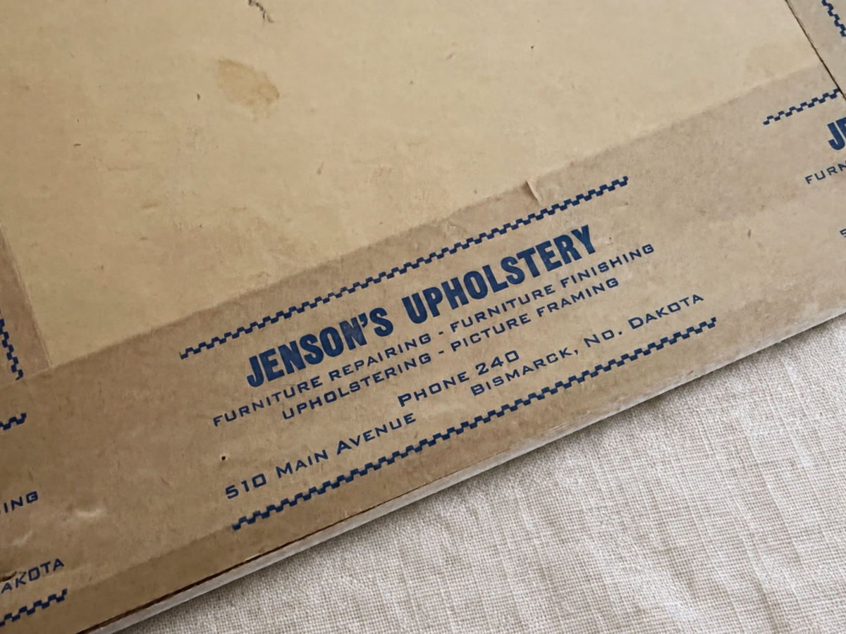 Close-up of blue Jenson’s Upholstery printing on brown framing tape. The label lists furniture repairing, furniture finishing, upholstering, picture framing, Phone 240, and 510 Main Avenue in Bismarck, North Dakota, with backing discoloration visible above.