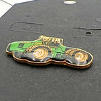 Close-up angled view of John Deere tractor enamel pin showing glossy green body, yellow wheels and gold edging, farming themed lapel pin for hats, jackets or bags, vintage agricultural accessory for tractor lovers and collectors