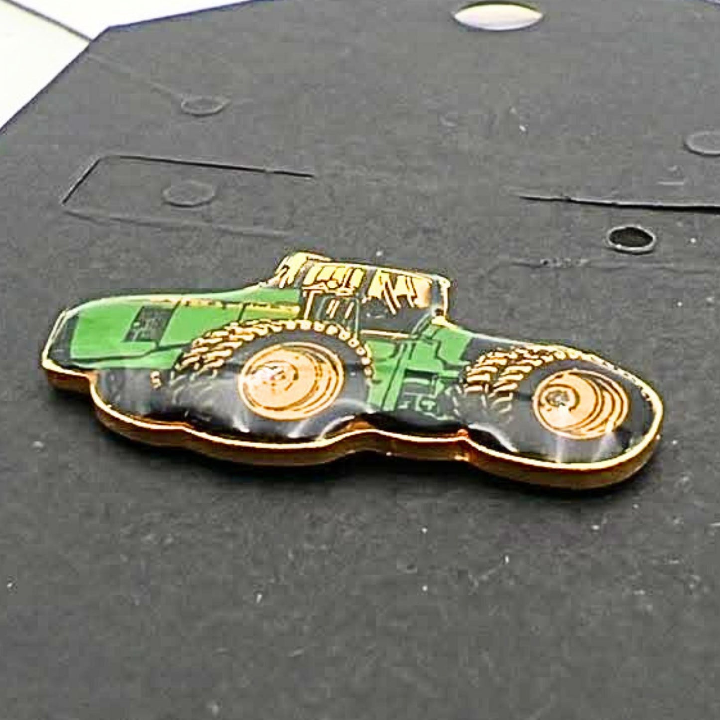 Close-up angled view of John Deere tractor enamel pin showing glossy green body, yellow wheels and gold edging, farming themed lapel pin for hats, jackets or bags, vintage agricultural accessory for tractor lovers and collectors