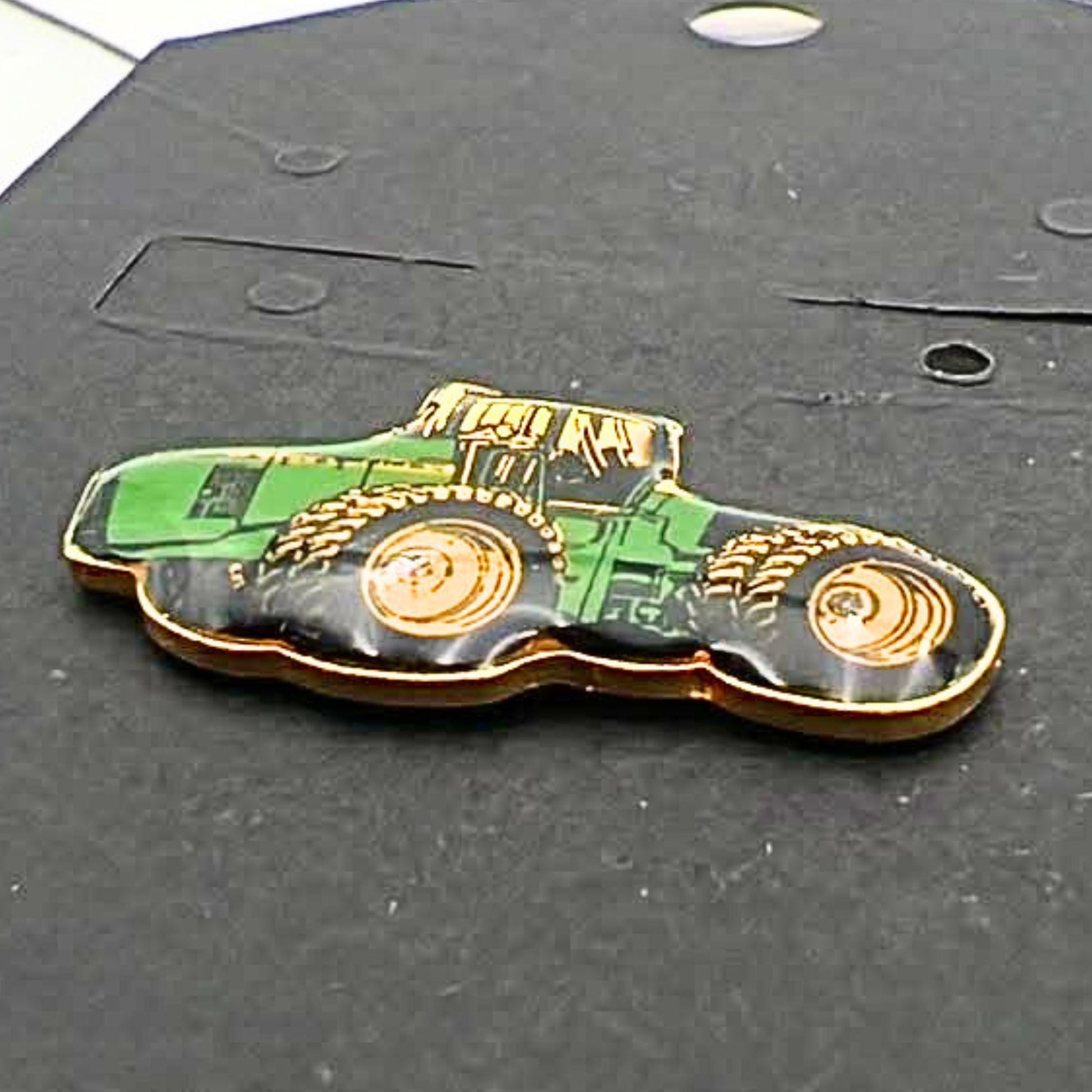 Close-up angled view of John Deere tractor enamel pin showing glossy green body, yellow wheels and gold edging, farming themed lapel pin for hats, jackets or bags, vintage agricultural accessory for tractor lovers and collectors
