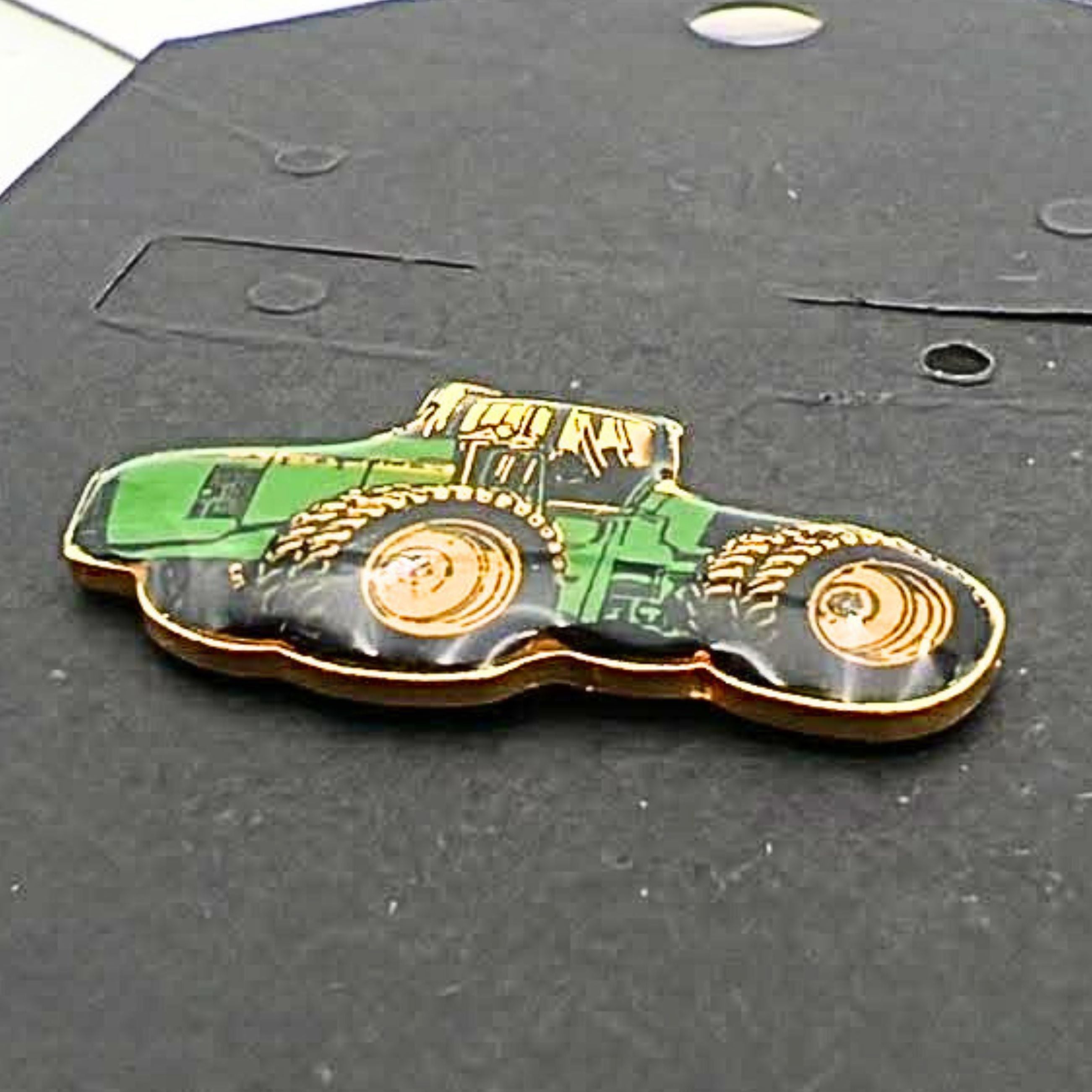 Close-up angled view of John Deere tractor enamel pin showing glossy green body, yellow wheels and gold edging, farming themed lapel pin for hats, jackets or bags, vintage agricultural accessory for tractor lovers and collectors