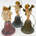 Angled perspective of vintage angel bell ornaments set featuring three musical cherubs on glitter bells. Instruments include cello, violin, and horn. Mid century West Germany Christmas decor with dimensional styling and vintage charm