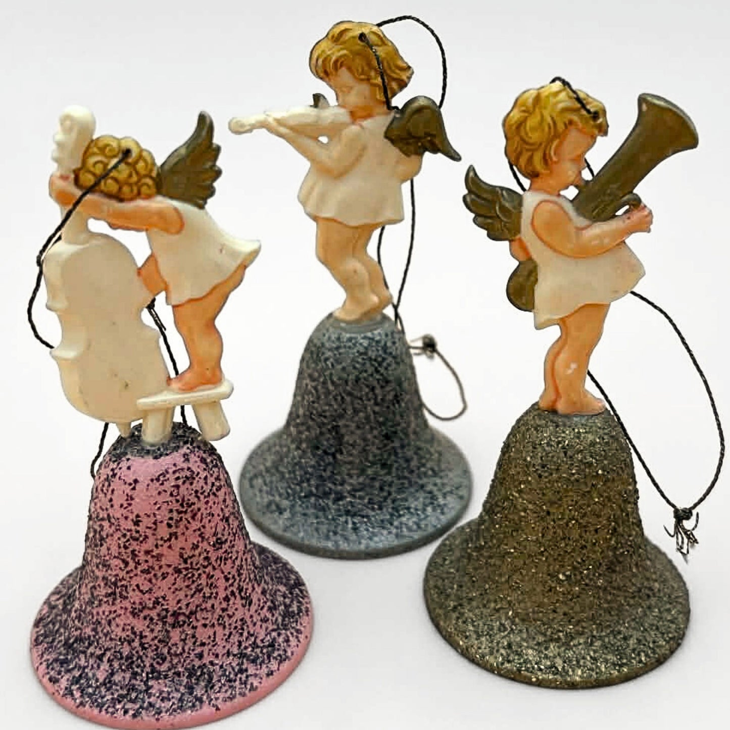 Angled perspective of vintage angel bell ornaments set featuring three musical cherubs on glitter bells. Instruments include cello, violin, and horn. Mid century West Germany Christmas decor with dimensional styling and vintage charm