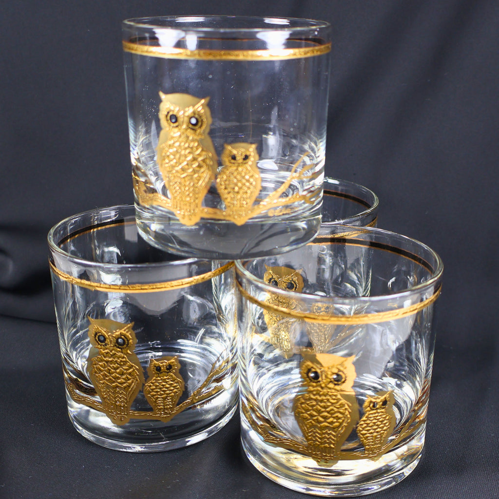 Complete set of four vintage Culver Ltd. owl rocks glasses displayed in a tiered arrangement against a charcoal background. Each clear tumbler features paired gold owls, dark eye details, a branching perch, and a coordinating metallic rim.