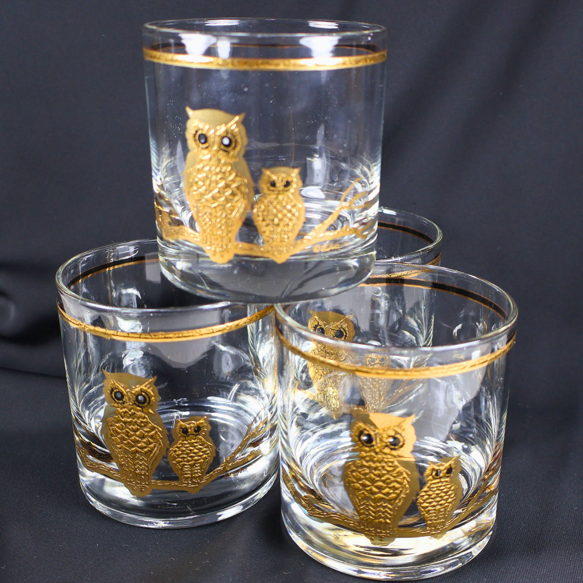 Complete set of four vintage Culver Ltd. owl rocks glasses displayed in a tiered arrangement against a charcoal background. Each clear tumbler features paired gold owls, dark eye details, a branching perch, and a coordinating metallic rim.