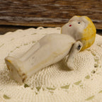 Side and underside view of the miniature bisque doll resting on an ivory crocheted textile. The photograph shows the sculpted profile, painted facial details, molded yellow hair, stationary joined legs, matte porcelain surface, and one movable arm at the shoulder.
