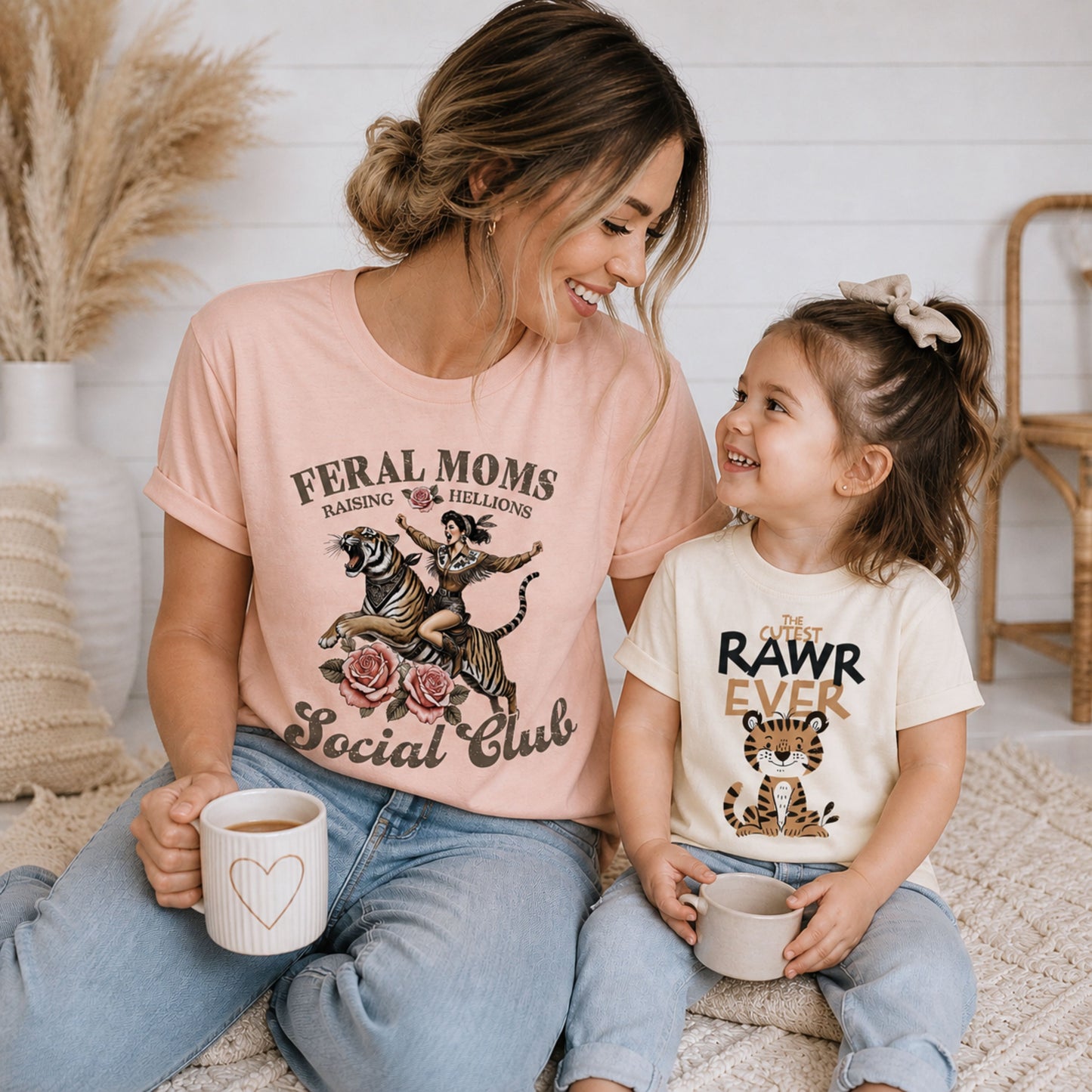 Smiling mom and toddler girl wearing coordinating tiger graphic tees, including “Feral Moms Social Club” and “Cutest RAWR Ever,” styled in a cozy neutral indoor lifestyle scene.