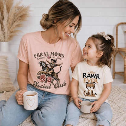 Smiling mom and toddler girl wearing coordinating tiger graphic tees, including “Feral Moms Social Club” and “Cutest RAWR Ever,” styled in a cozy neutral indoor lifestyle scene.