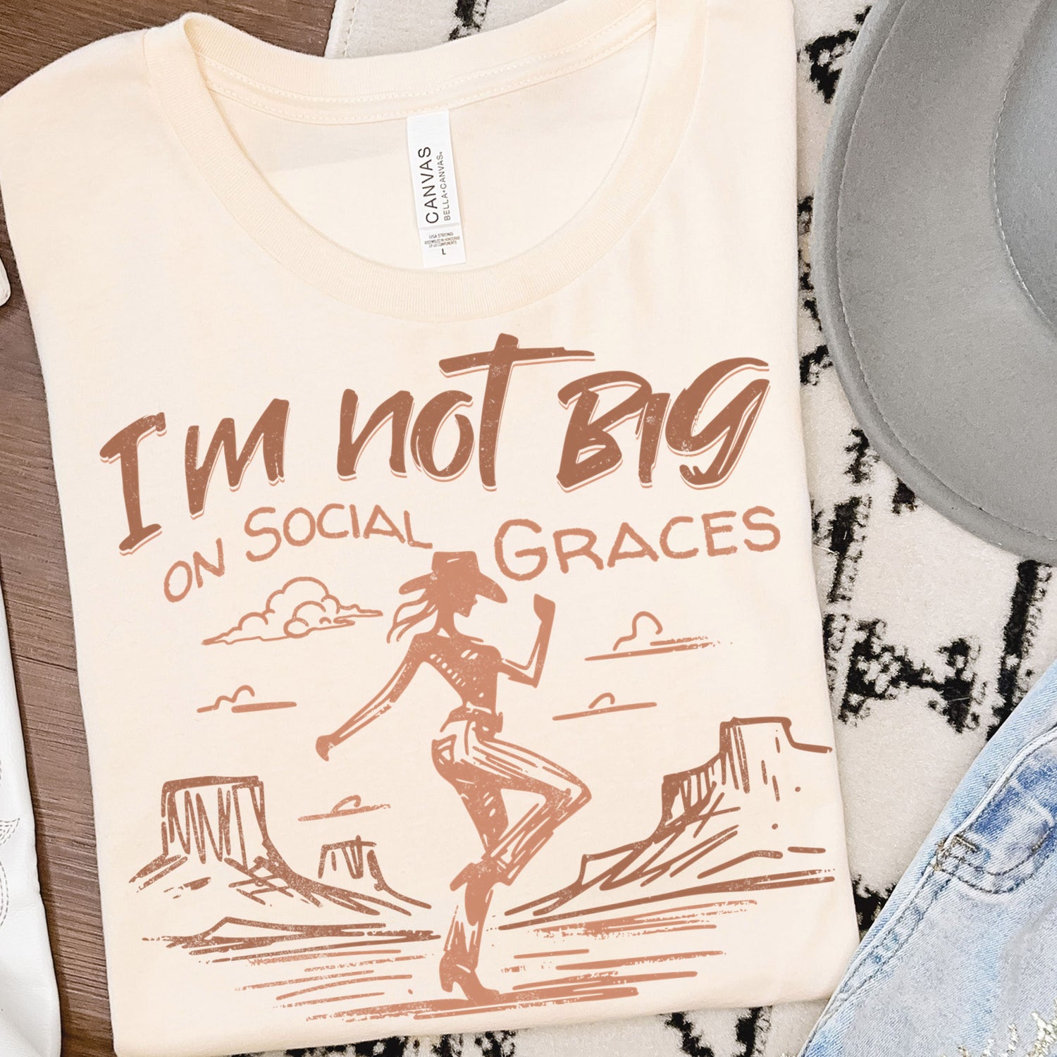 Neutral cream western graphic tee folded in flat lay with cowgirl design and desert illustration, boutique t-shirt styled with western accessories