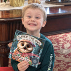 A smiling child in a teal shirt holds Oliver the Owl: A Sweet Season with its illustrated holiday cover facing the camera, seated beside a wooden piano decorated for Christmas.
