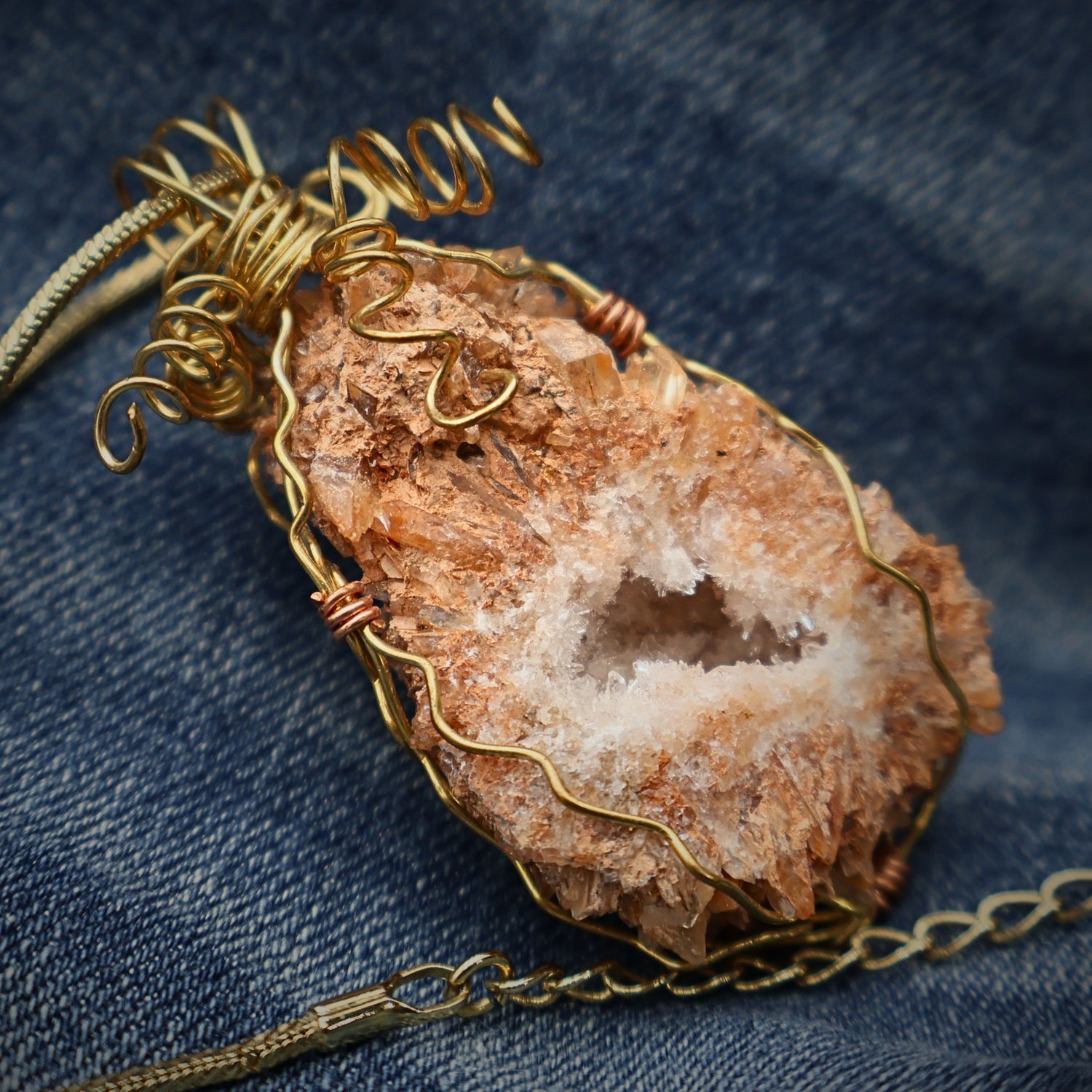 Peach druzy quartz geode necklace with gold wire wrapped pendant resting on denim fabric, showcasing raw crystal texture and natural stone sparkle, boho western statement jewelry piece with earthy tones, unique handmade crystal necklace for women gift idea
