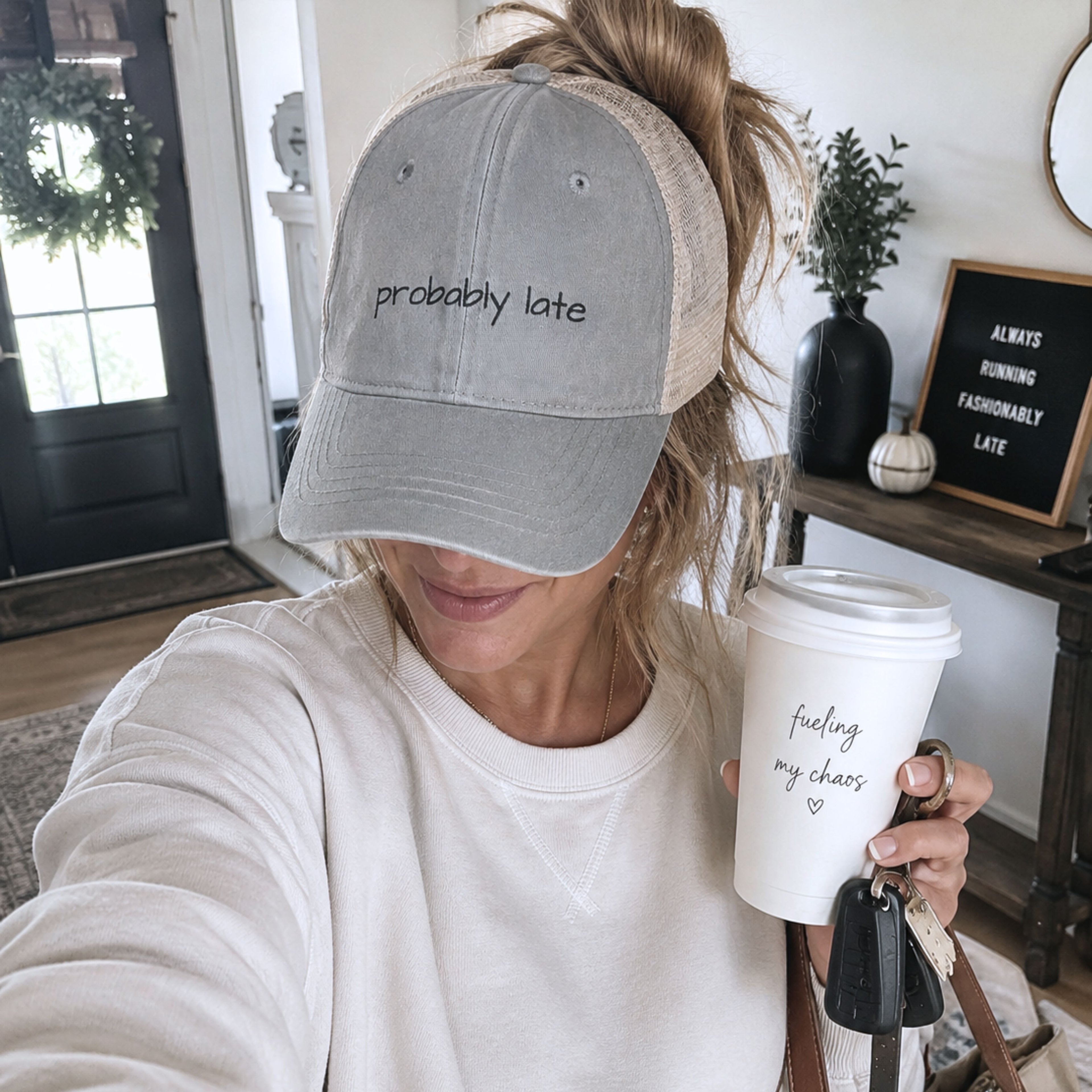 Woman wearing gray and tan mesh back trucker hat with probably late embroidered saying while holding coffee and keys in a casual lifestyle selfie