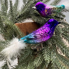 Purple glass bird ornament in the foreground, showing its blue-green rear body, silver-glitter wing decoration, white feather tail, and spring-mounted metal clip. A coordinating green and purple bird sits above it among evergreen branches beside wood and cream lace.