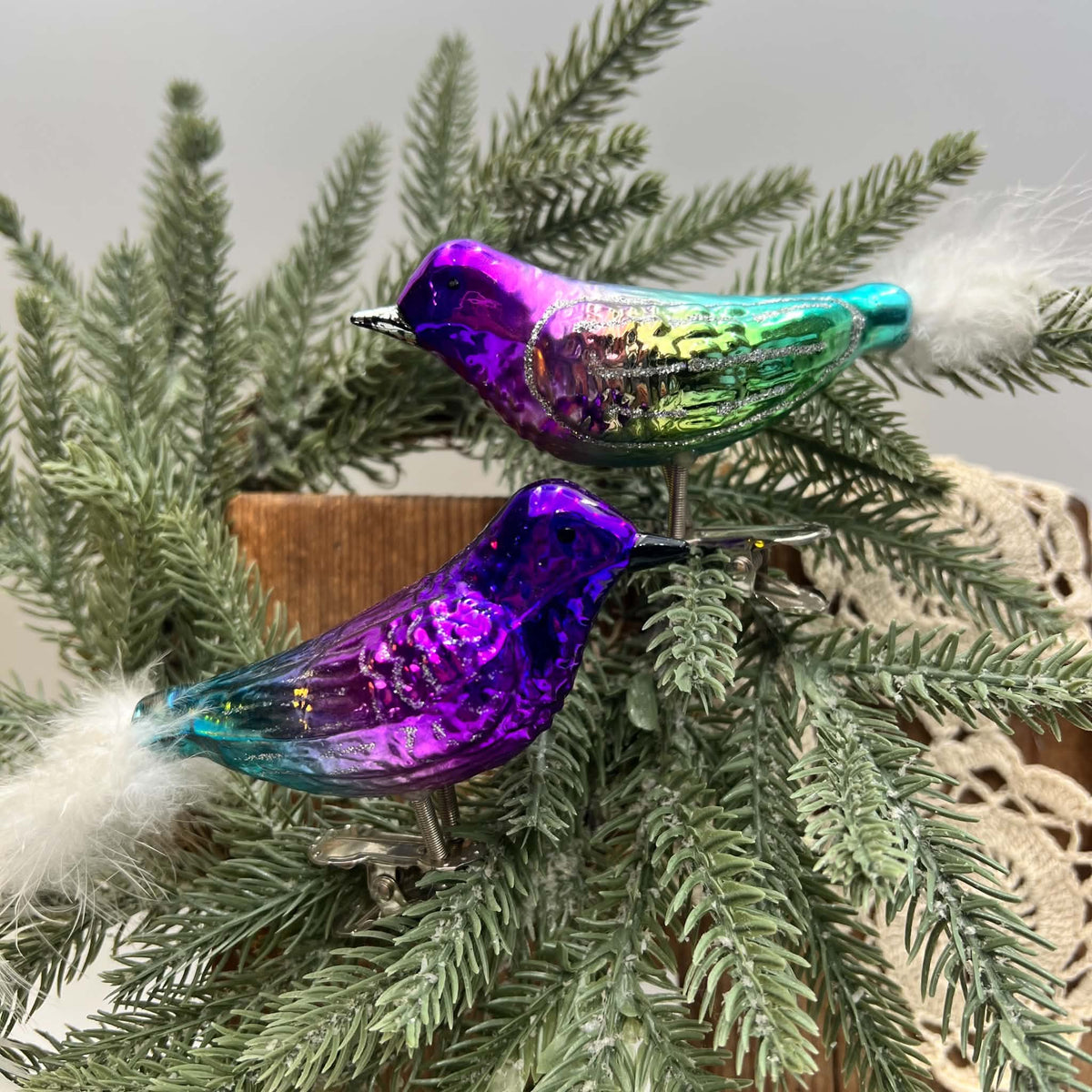 Pair of purple and teal glass bird Christmas ornaments perched among evergreen branches, with white feather tails and silver metal clips. The upper bird has green and silver wing coloring, while the lower bird shows deeper violet and blue beside a wooden backdrop.
