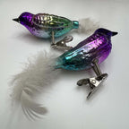 Angled view of a coordinating pair of purple and turquoise glass bird ornaments on white. Fluffy white feather tails, dark pointed beaks, silver-glitter wing accents, and shiny metal branch clips are visible, with the violet bird closer to the camera than the green bird.