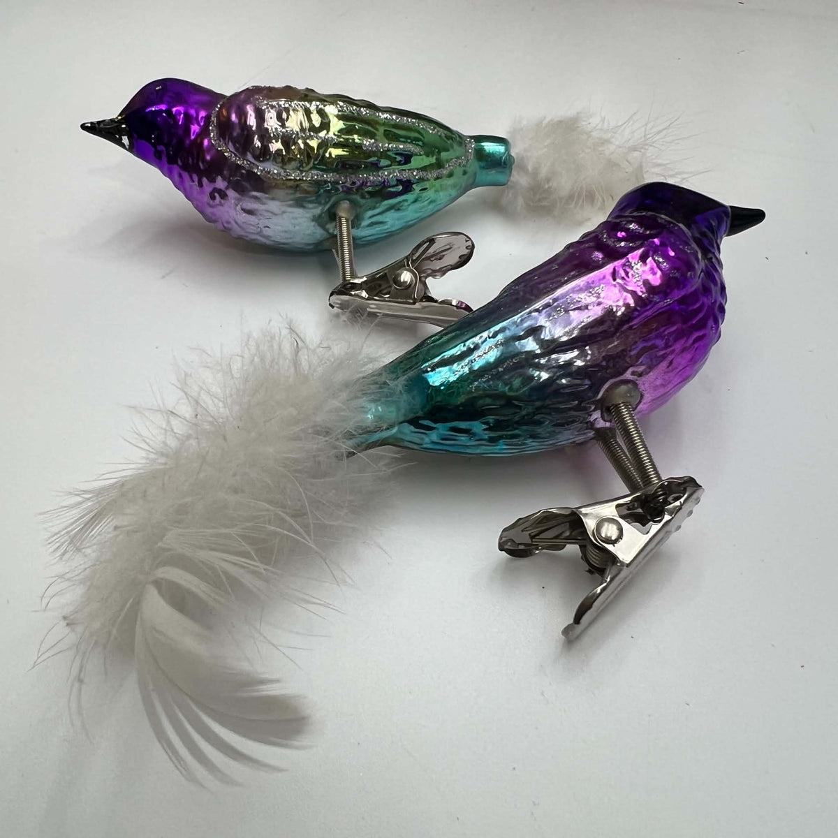 Angled view of a coordinating pair of purple and turquoise glass bird ornaments on white. Fluffy white feather tails, dark pointed beaks, silver-glitter wing accents, and shiny metal branch clips are visible, with the violet bird closer to the camera than the green bird.
