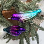 Overhead view of two reflective glass bird ornaments arranged on evergreen branches over a wooden board and cream lace. One bird blends purple into silver, green, and turquoise; the other has a darker violet body. White feather tails extend in opposite directions.