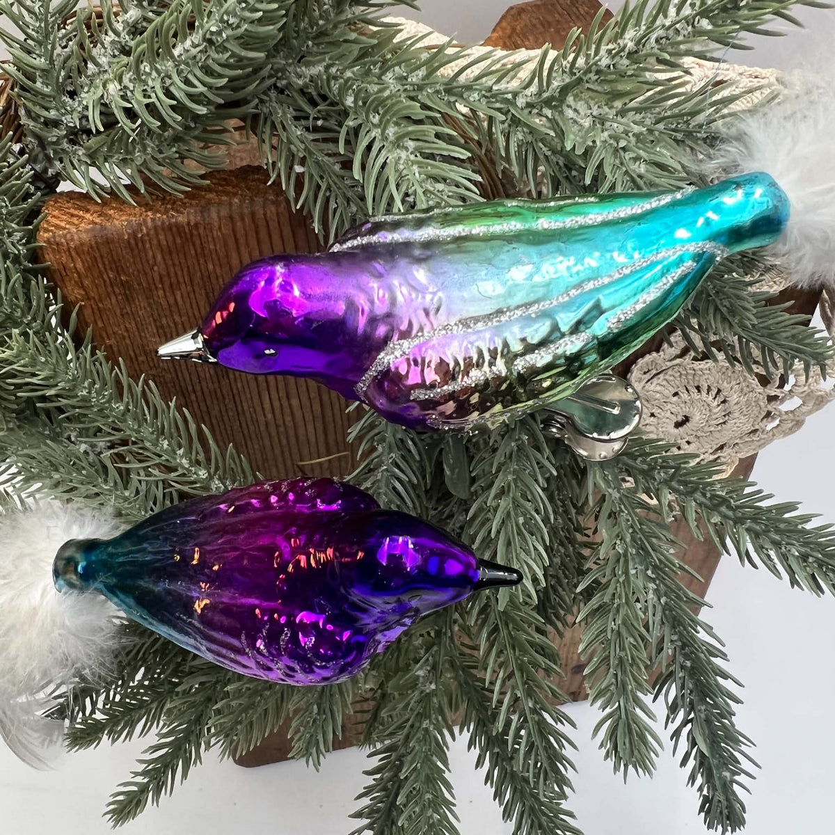 Overhead view of two reflective glass bird ornaments arranged on evergreen branches over a wooden board and cream lace. One bird blends purple into silver, green, and turquoise; the other has a darker violet body. White feather tails extend in opposite directions.