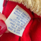 Detail of the vintage Santa doll’s sewn cloth manufacturer label, showing blue Sakai & Co., Ltd., Tokyo Japan, and Made in Japan lettering beneath removal-warning text. A hand with red nail polish holds the label against the red suit below yellowed fuzzy trim.