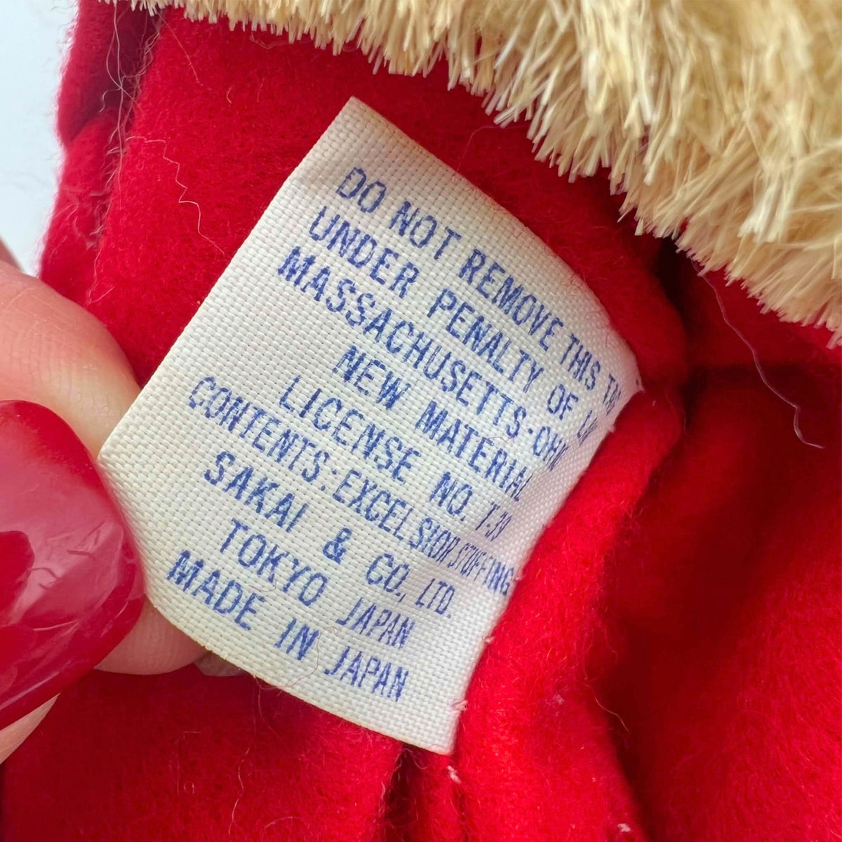 Detail of the vintage Santa doll’s sewn cloth manufacturer label, showing blue Sakai & Co., Ltd., Tokyo Japan, and Made in Japan lettering beneath removal-warning text. A hand with red nail polish holds the label against the red suit below yellowed fuzzy trim.