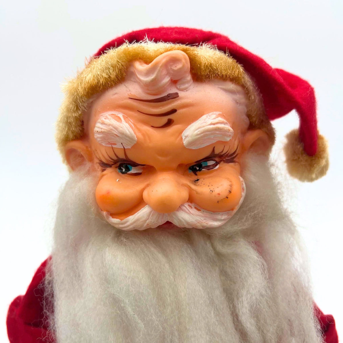 Close-up of the vintage Santa doll’s molded face, showing blue sideways-looking eyes, white painted eyebrows, a curled mustache, and a long fiber beard. Dark marks and paint wear are visible on the cheeks beneath a red cap edged with yellowed fuzzy trim.