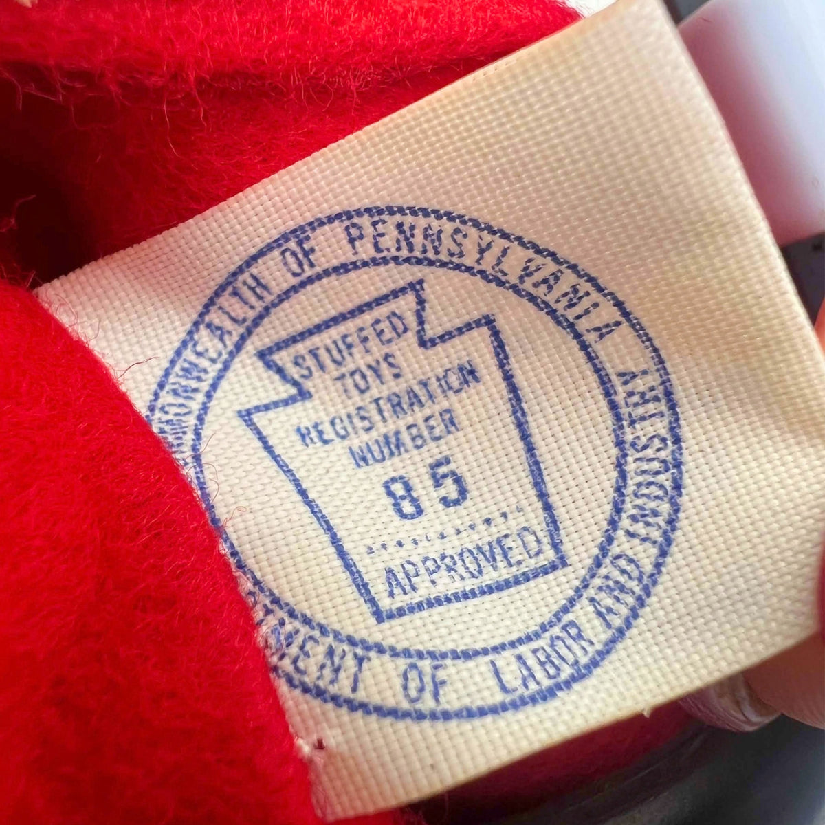 Close-up of a cream cloth label on the vintage Santa doll, printed in blue with a Commonwealth of Pennsylvania stuffed-toy approval stamp and registration number 85. Red fabric surrounds the textured tag, and a hand holds it open beside part of the metal stand.