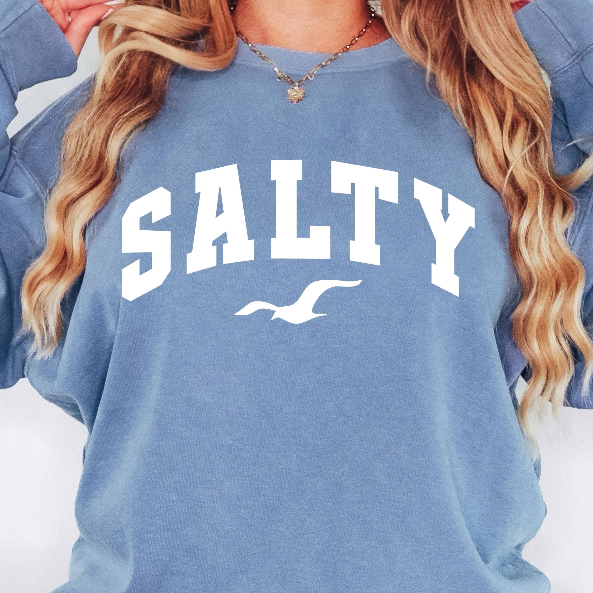 Woman wearing blue jean SALTY sweatshirt, oversized beach crewneck, relaxed coastal outfit, casual seaside fashion, comfortable vacation clothing for women, nautical graphic sweatshirt