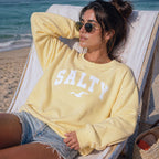 Woman wearing butter yellow SALTY sweatshirt lounging in beach chair, sunny coastal setting, relaxed vacation outfit, oversized crewneck sweatshirt, comfortable beachwear for women, soft pastel sweatshirt, seaside casual style with ocean backdrop
