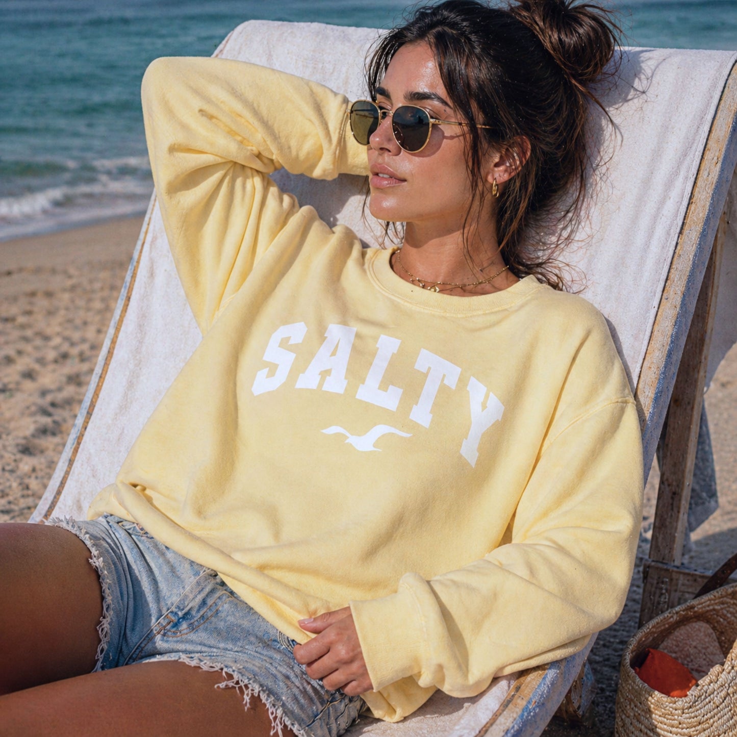 Woman wearing butter yellow SALTY sweatshirt lounging in beach chair, sunny coastal setting, relaxed vacation outfit, oversized crewneck sweatshirt, comfortable beachwear for women, soft pastel sweatshirt, seaside casual style with ocean backdrop