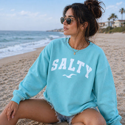 Woman wearing chalky mint SALTY sweatshirt sitting on beach, relaxed coastal outfit, oversized crewneck sweatshirt, comfortable vacation clothing, beachwear for women, nautical graphic sweatshirt, seaside casual style