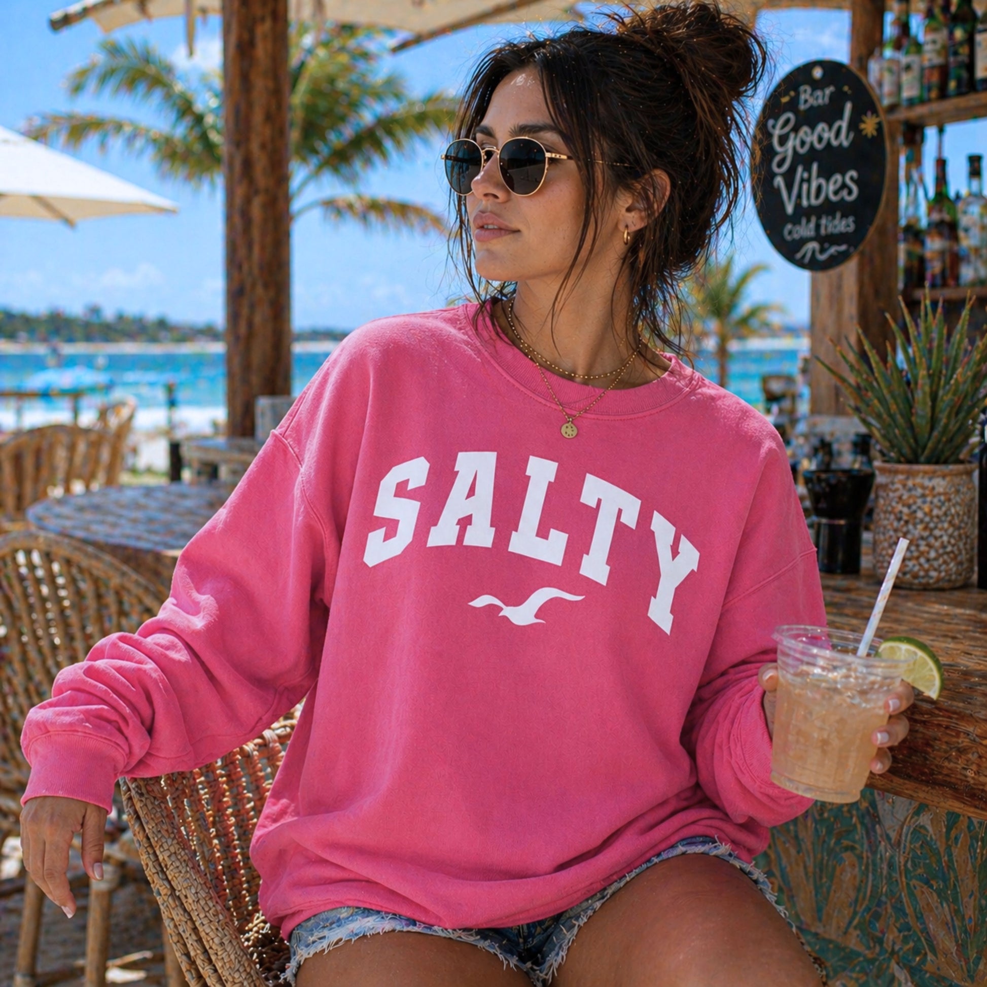 Woman wearing crimson SALTY sweatshirt at beach resort setting, bold coastal outfit, oversized crewneck sweatshirt, relaxed vacation style clothing, nautical graphic sweatshirt, trendy beachwear for women, sunny seaside lifestyle
