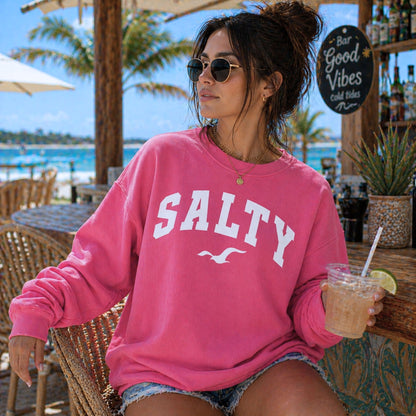 Woman wearing crimson SALTY sweatshirt at beach resort setting, bold coastal outfit, oversized crewneck sweatshirt, relaxed vacation style clothing, nautical graphic sweatshirt, trendy beachwear for women, sunny seaside lifestyle