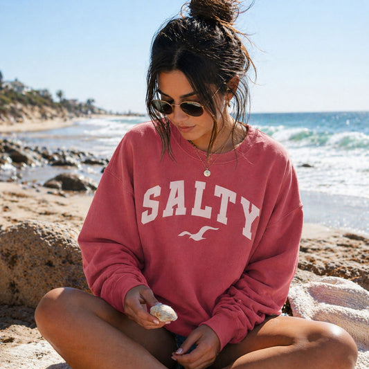 Woman wearing crimson SALTY beach sweatshirt sitting on sandy beach looking at seashell, relaxed coastal lifestyle outfit, oversized graphic crewneck, ocean background, vacation style outfit, comfy beachwear for women, nautical aesthetic sweatshirt