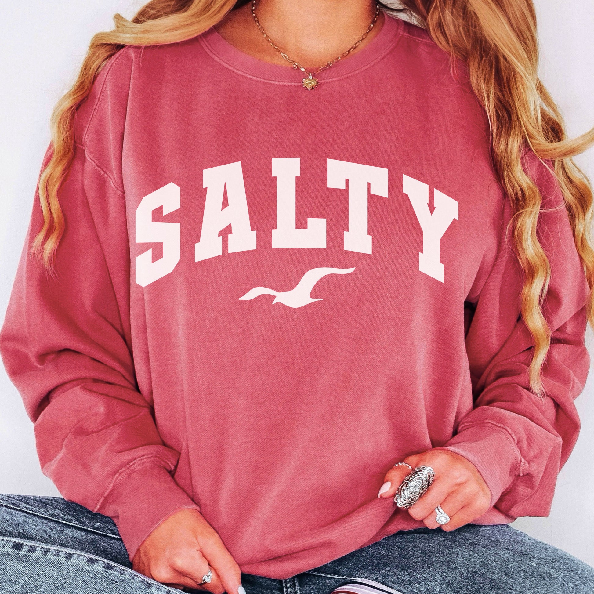 Woman wearing crimson SALTY beach sweatshirt, oversized coastal crewneck, relaxed vacation outfit for women, casual seaside fashion, soft cotton blend sweatshirt, nautical graphic top, beach lifestyle clothing