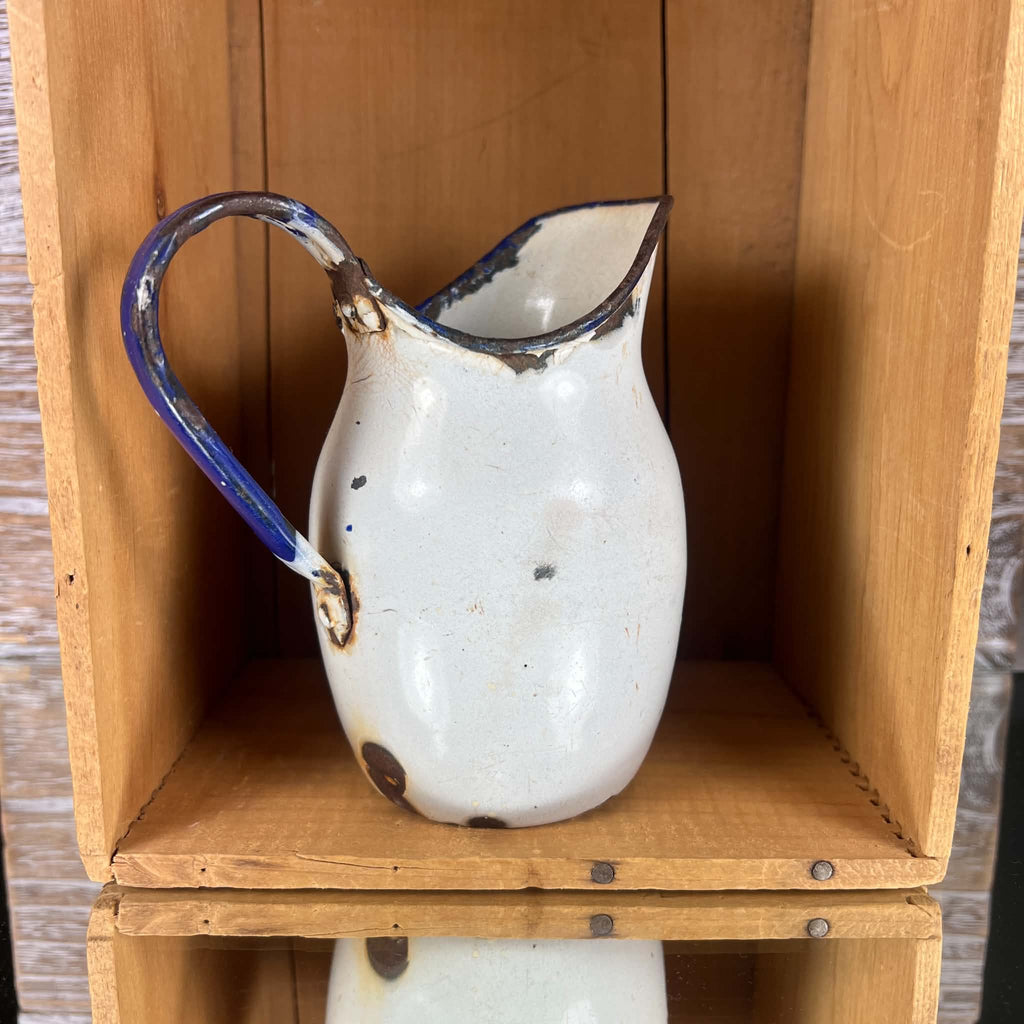White and cobalt-blue enamelware pitcher photographed from behind inside a wooden crate. The curved handle arches above the opening, with extensive coating loss around its attachments and irregular brown patches across the rounded body and near the bottom edge.