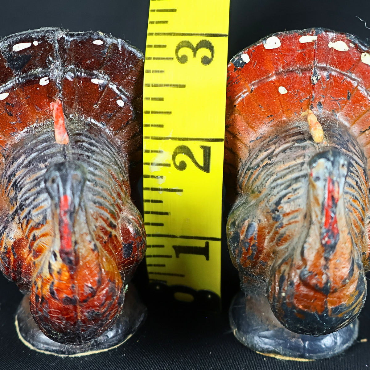 Pair of vintage Gurley turkey candles photographed beside a yellow measuring tape showing a height of approximately three inches. Their broad feathered tails, rounded bodies, painted red facial details, white wax losses, upright wicks and aged surfaces are clearly visible.