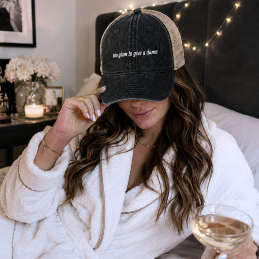Woman in a white robe wearing too glam to give a damn embroidered ponytail trucker hat holding a glass of wine