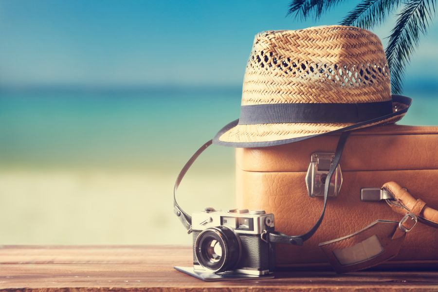 Vintage travel scene with a camera, leather suitcase, and straw hat by the beach, capturing a nostalgic summer travel vibe and used as a background image for a travel and souvenir collection.