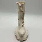 Front-facing view of a vintage ceramic high-button boot planter standing on a decorative scrolling pedestal. The narrow profile, pointed toe, centered heel and raised trim can be seen clearly, along with the cream finish and brown shading used to define the molding.