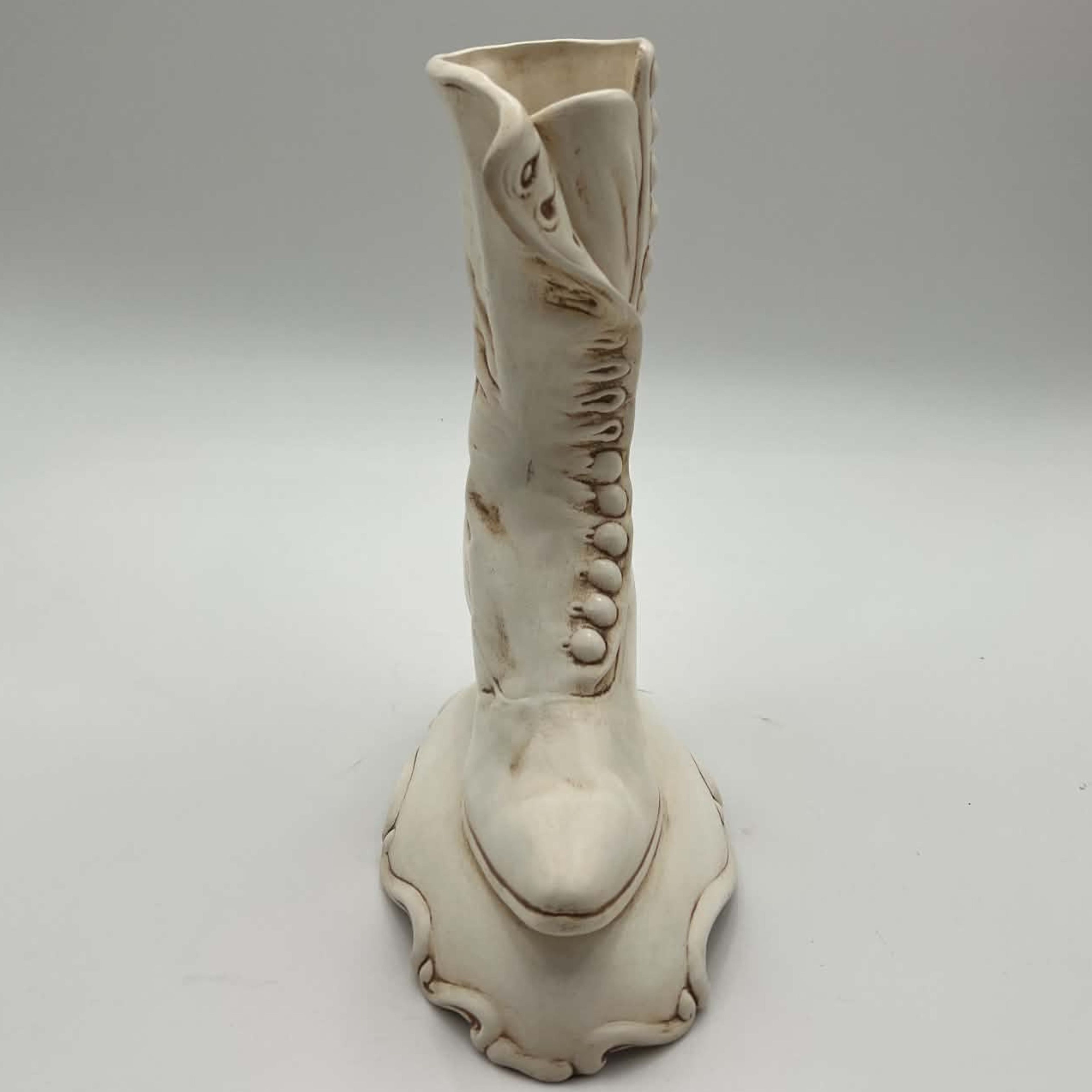 Front-facing view of a vintage ceramic high-button boot planter standing on a decorative scrolling pedestal. The narrow profile, pointed toe, centered heel and raised trim can be seen clearly, along with the cream finish and brown shading used to define the molding.